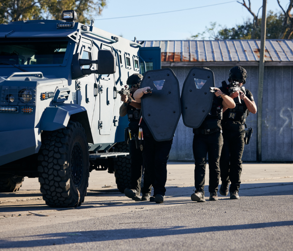 A group of uniformed St. Augustine Police Officers with the Special Response Team (SRT) stacked in formation next to an armored vehicle. Two officers in the front have ballistic shields with their handguns drawn.