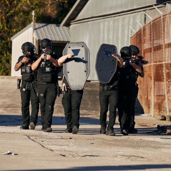 A group of uniformed St. Augustine Police Officers with the Special Response Team (SRT) stacked in formation next to an armored vehicle. Two officers in the front have ballistic shields with their handguns drawn.